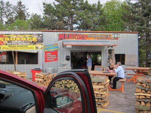 A rustic BBQ restaurant with a sign reading "Meaty's Roadhouse BBQ," surrounded by stacked firewood and patrons outside.