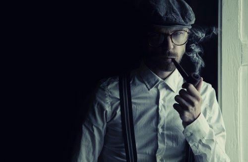 A man in a cap and glasses smokes a pipe, dressed in a white shirt and suspenders, standing by a window.