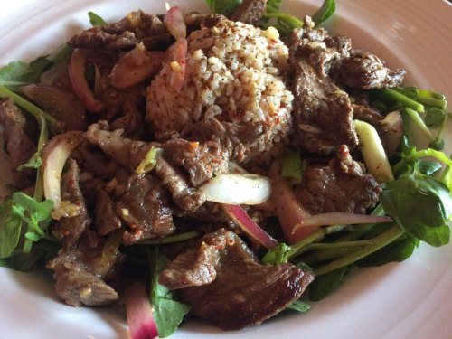 A plate of seasoned beef slices on a bed of greens, topped with onions and served with a mound of rice.