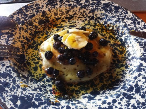 A stack of pancakes topped with blueberries and banana slices, served on a speckled blue plate with syrup.