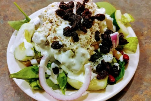 A colorful salad topped with shredded chicken, yogurt dressing, raisins, and various fresh vegetables on a plate.