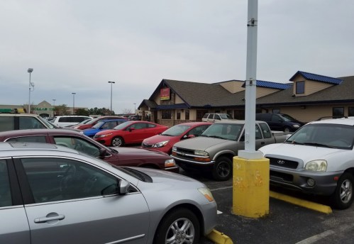 A busy parking lot filled with various cars in front of a restaurant under a cloudy sky.