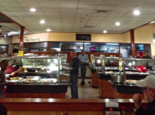 A busy buffet restaurant with diners serving themselves from various food stations under bright lights.