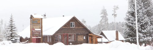 A rustic wooden cabin in a snowy landscape, surrounded by tall trees and a sign for a nearby lodge.