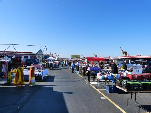 A bustling outdoor market with colorful stalls, shoppers browsing, and clear blue skies overhead.