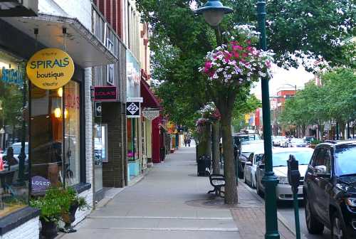 A charming street lined with shops, trees, and colorful flower baskets, with parked cars and a bench visible.