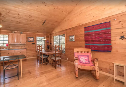 Cozy wooden interior of a cabin featuring a dining area, kitchen, and a colorful wall hanging.