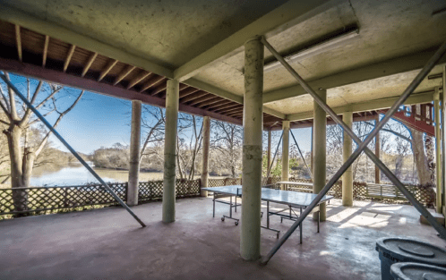 An open space with concrete pillars, overlooking a river and trees, featuring a table and a rustic railing.