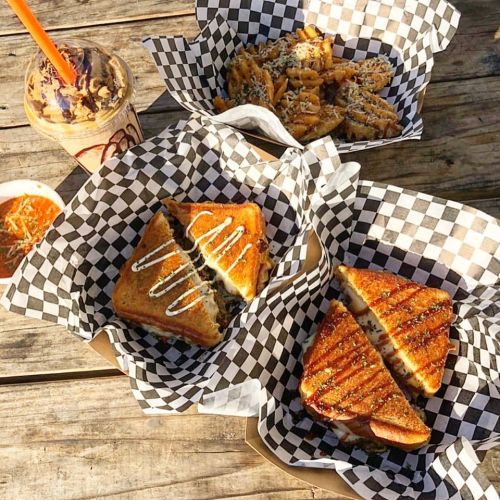 A table with three checkered paper trays: two grilled sandwiches, a serving of fries, and a drink.