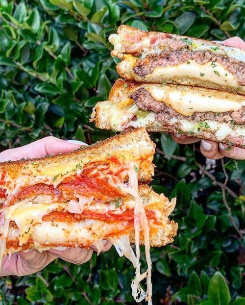 Two hands hold stacked sandwiches filled with melted cheese, pepperoni, and meat, against a backdrop of green leaves.