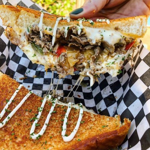 A hand holds a cheesy sandwich filled with beef, veggies, and drizzled with sauce, resting on a checkered paper.