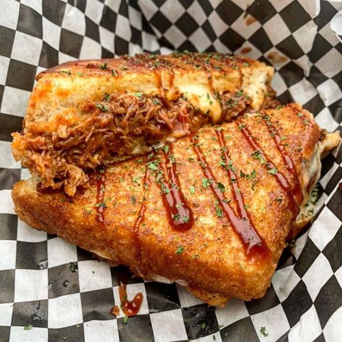 Grilled cheese sandwich with pulled pork and barbecue sauce, served in a checkered paper basket.