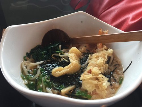 A bowl of noodles topped with seaweed, green onions, and tempura, with a wooden spoon resting inside.