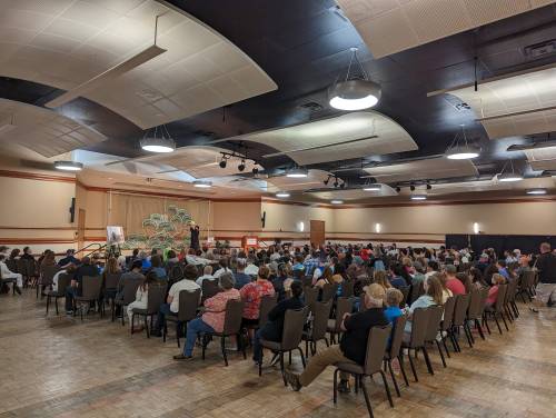 A large audience seated in a hall, facing a speaker at a podium with decorative plants in the background.
