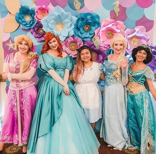 Five women dressed as princesses stand in front of a colorful flower backdrop, smiling and posing for the photo.