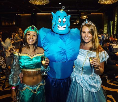 Two women in princess costumes pose with a man in a blue genie costume, all holding drinks at a festive event.