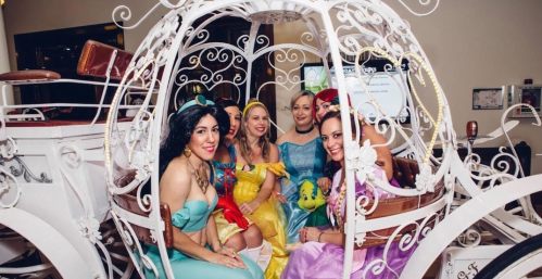 Five women in colorful princess costumes sitting inside a decorative white carriage.