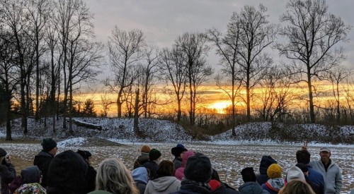 A crowd of people watches a sunset behind bare trees in a snowy landscape.
