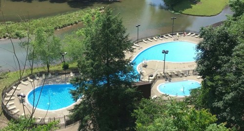 Aerial view of two blue swimming pools surrounded by greenery and a river, with lounge chairs nearby.
