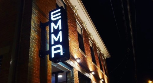 A brightly lit sign reading "EMMA" on a brick building at night.