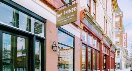 Exterior of a shop named "The Rhined," featuring a sign for artisanal cheese and provisions, with large windows.