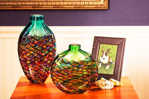 Two colorful glass vases with spiral designs on a wooden table, alongside a framed photo of a dog and a seashell.