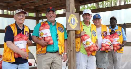 Five men in yellow vests hold bags of apples, standing under a wooden shelter with a Lions Club emblem.