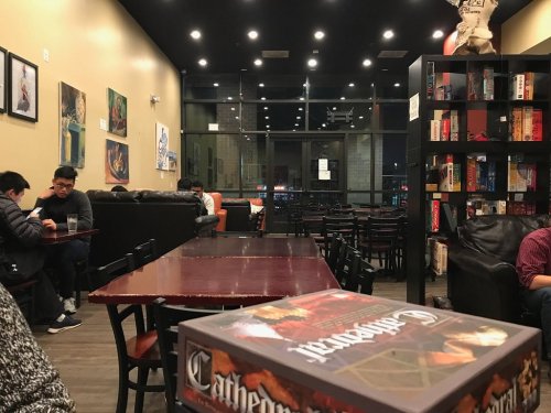 A cozy café interior with dark walls, tables, and a bookshelf filled with board games, patrons enjoying their time.