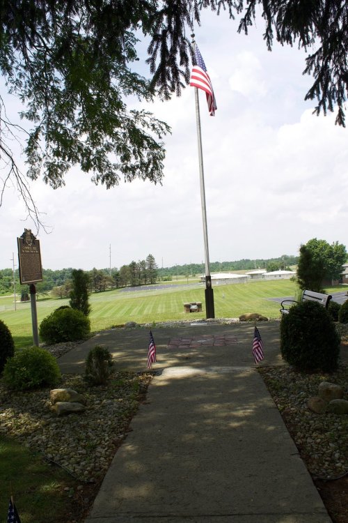 A pathway leads to a flagpole with an American flag, surrounded by greenery and small flags, overlooking a field.
