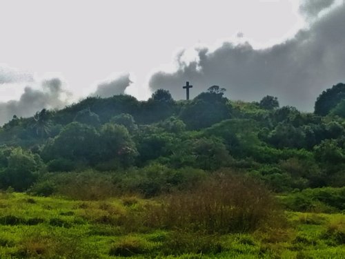 A cross stands atop a green hill, surrounded by lush vegetation and a cloudy sky.