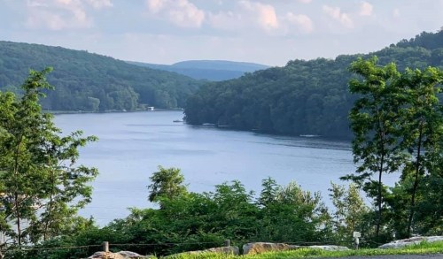 A serene lake surrounded by lush green trees and rolling hills under a partly cloudy sky.