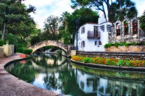 A serene canal scene with a stone bridge, colorful flowers, and charming buildings surrounded by lush greenery.
