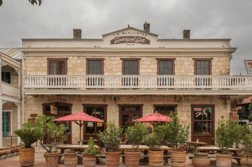 Historic stone building with a balcony, featuring outdoor seating and red umbrellas, under a cloudy sky.