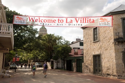 A banner reading "Welcome to La Villita" hangs over a street with shops and trees in a historic area.
