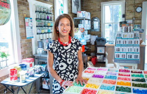 A smiling woman in a patterned dress stands behind a table filled with colorful wax melts in a cozy shop.