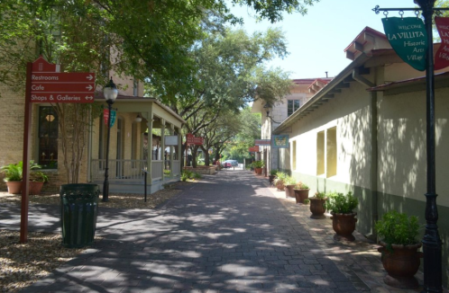 A charming street lined with shops and greenery, featuring directional signs and a welcoming atmosphere.