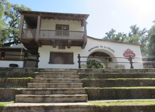A white building with a balcony and stairs, labeled "Entrance to La Villita," surrounded by greenery.