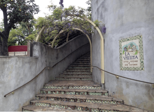 A stone staircase leads up to an archway adorned with purple flowers, with a sign reading "La Villita, Little Village."