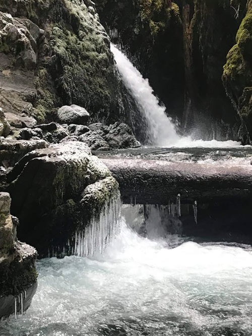 A serene waterfall cascades over moss-covered rocks into a clear, flowing river, surrounded by lush greenery.