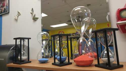 A row of colorful hourglasses on a wooden shelf, with a mirror and vibrant wall art in the background.
