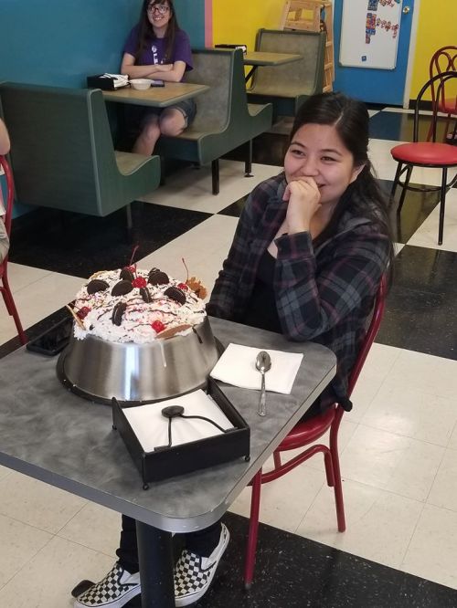 A young woman smiles while sitting at a table with a large ice cream cake in front of her. Colorful diner background.