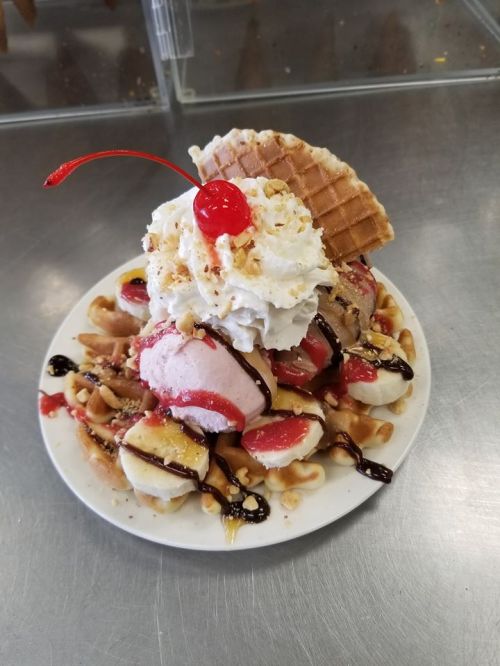 A colorful ice cream sundae topped with whipped cream, a cherry, and a waffle, served on a plate with banana and chocolate sauce.