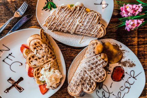 Two plates of beautifully presented pancakes with fruit, whipped cream, and drizzled sauces on a wooden table.