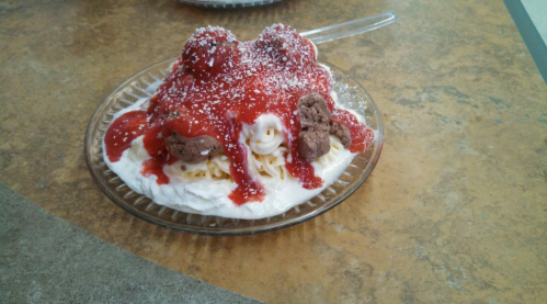 A dessert with whipped cream, chocolate pieces, and strawberry sauce, topped with powdered sugar on a glass plate.