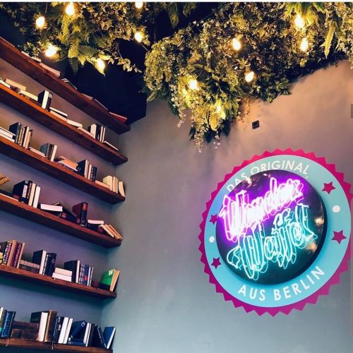 A cozy café interior featuring a neon sign, bookshelves, and hanging greenery.