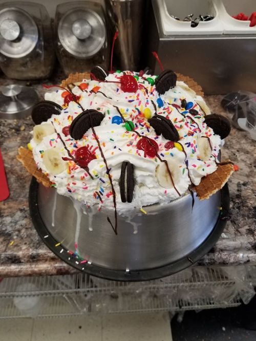 A large, colorful ice cream cake topped with whipped cream, cherries, Oreos, sprinkles, and banana slices.
