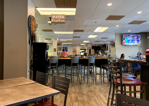 Interior of a casual dining space with a bar, tables, and a water station sign. People are seated in the background.