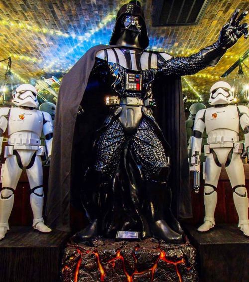 A life-sized Darth Vader statue stands between two Stormtrooper figures in a dimly lit, decorated space.