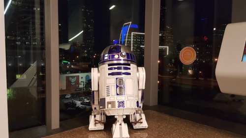 A robot resembling R2-D2 stands by a window, with a city skyline visible at night in the background.
