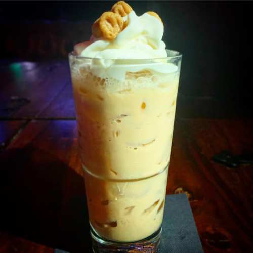 A tall glass filled with iced coffee topped with whipped cream and cereal pieces.